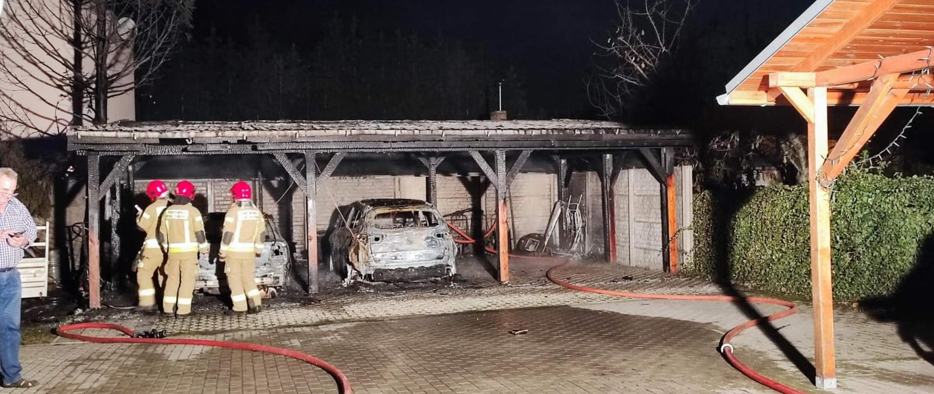Widać spaloną wiatę garażową, dwa samochody, jest ciemno
