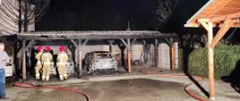 Widać spaloną wiatę garażową, dwa samochody, jest ciemno