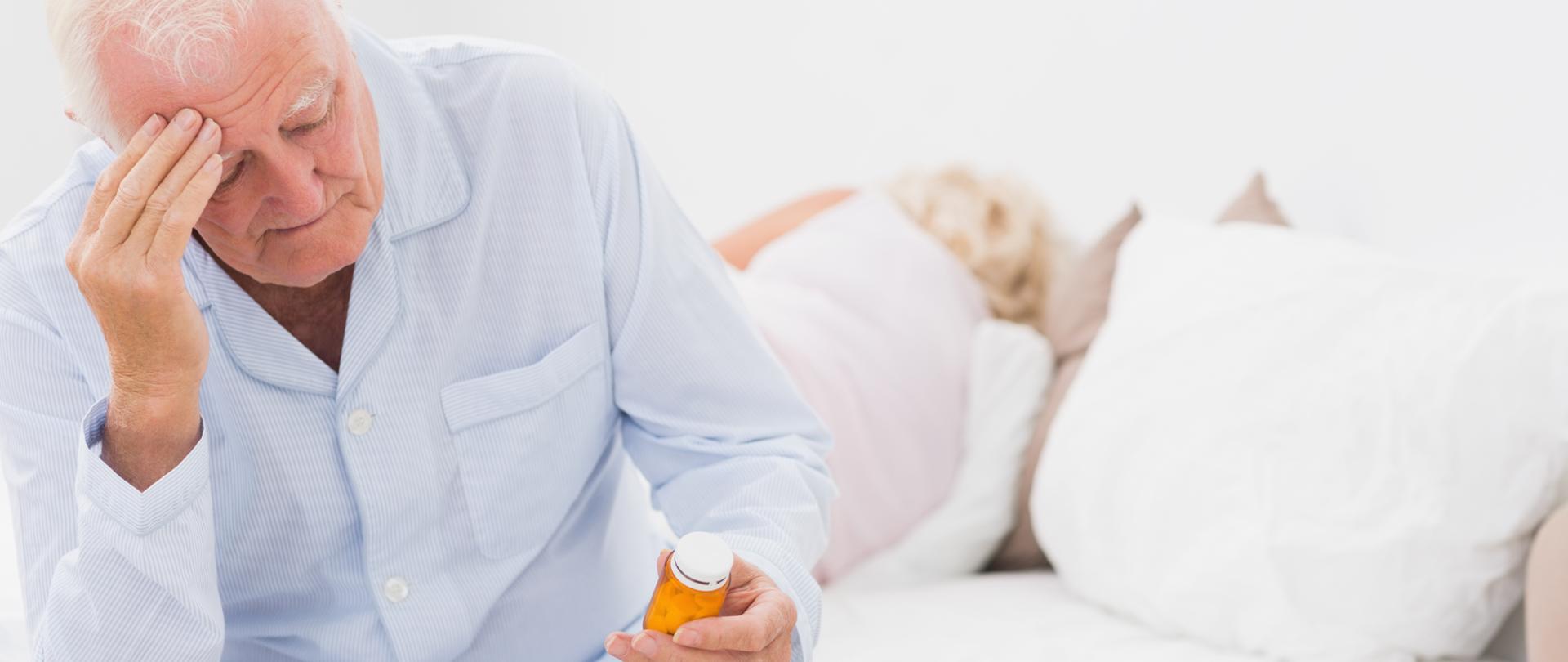 starsza kobieta śpi, a starszy mężczyzna siedzi na łóżku, trzyma się za głowę, a w ręku trzyma leki
