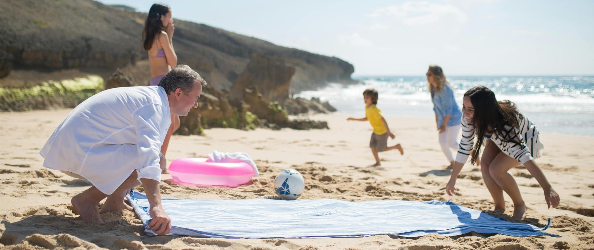  Na zdjęciu widzimy plażę z czterema osobami, których twarze są zamazane w celu zachowania prywatności. Dorosła osoba w białej koszuli i szortach układa na piasku niebiesko-białą ręcznik. Obok stoi inna dorosła osoba w bikini, patrząc w kierunku morza. W tle dwójka dzieci bawi się przy brzegu wody: jedno wydaje się biec w stronę oceanu, a drugie jest bliżej brzegu, prawdopodobnie zabawiając się z falami. Plaża jest słoneczna i przyjemna, z bezchmurnym niebem i spokojnym morzem, co czyni ją interesującym miejscem do rekreacji.