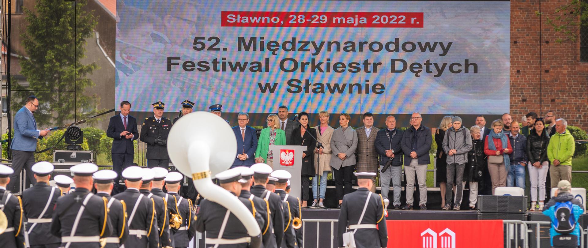 Na pierwszym planie stoją członkowie orkiestry dętej w mundurach galowych. W rękach trzymają instrumenty. Są odwróceni tyłem do fotografa. Na dalszym planie są zaproszeni goście oraz organizatorzy wydarzenia, którzy stoją na scenie.