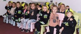 spora grupka dzieci w wieku przedszkolnym siedzi na kolorowych krzesełkach w sali prawie wszystkie dzieci mają na sobie dopasowane do nich ubranka strażackie w czarnym kolorze z odblaskami na głowach mają żółte hełmy
