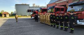 Strażacy stoją w pododdziale przed pojazdami pożarniczymi podczas uroczystej zbiórki. 