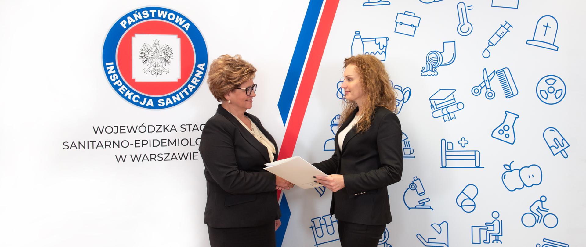 Dwie kobiety w strojach formalnych podają sobie ręce na tle ścianki z logo Państwowej Inspekcji Sanitarnej. Jedna trzyma w rękach teczkę z dokumentem powołania. 