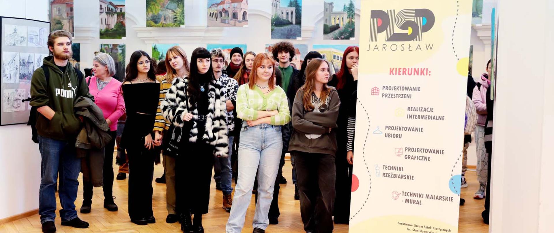 Zdjęcie przedstawia grupę młodzieży na tle prac malarskich. Po prawej stroni jest umieszczony pionowy baner reklamowy szkoły. 