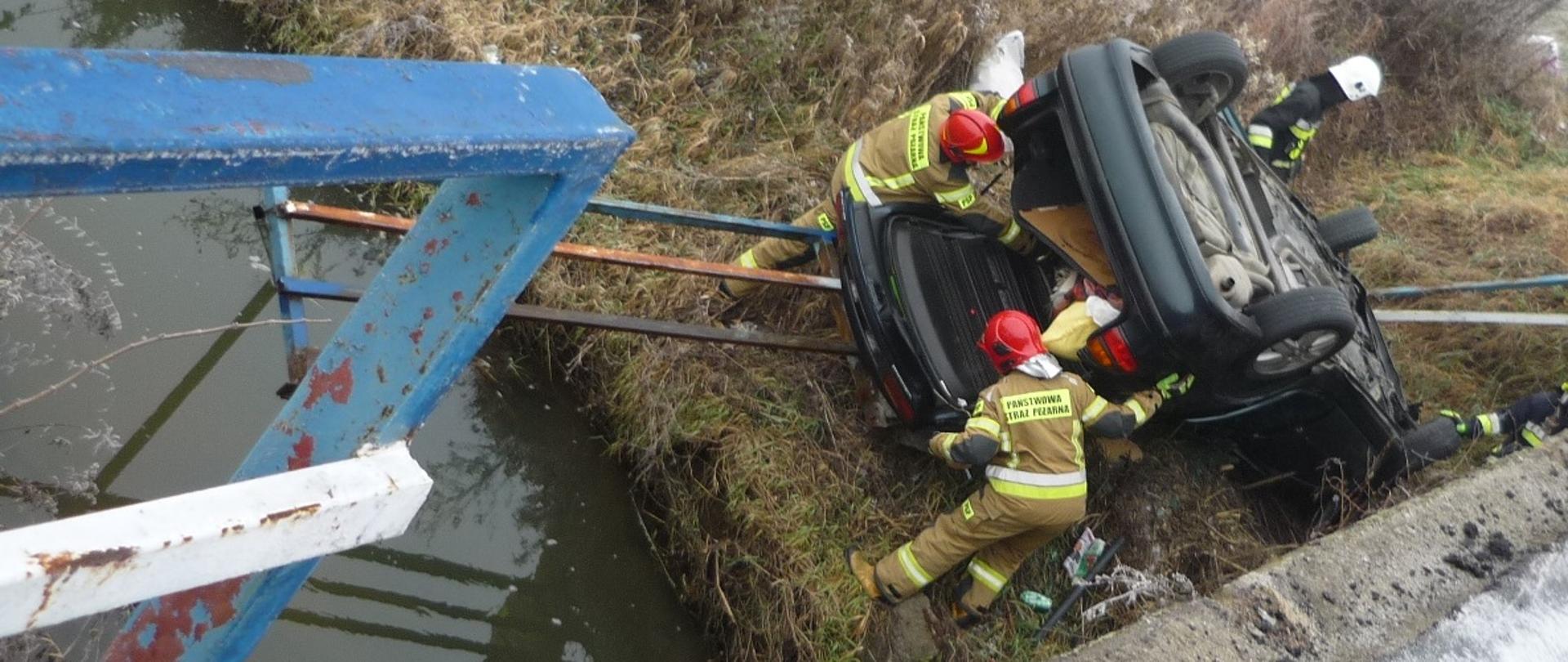 na zdjęciu widać samochód osobowy na dachu obok rzeki Żabnica, uszkodzoną bariere energochłonną mostu, oraz strażaków.