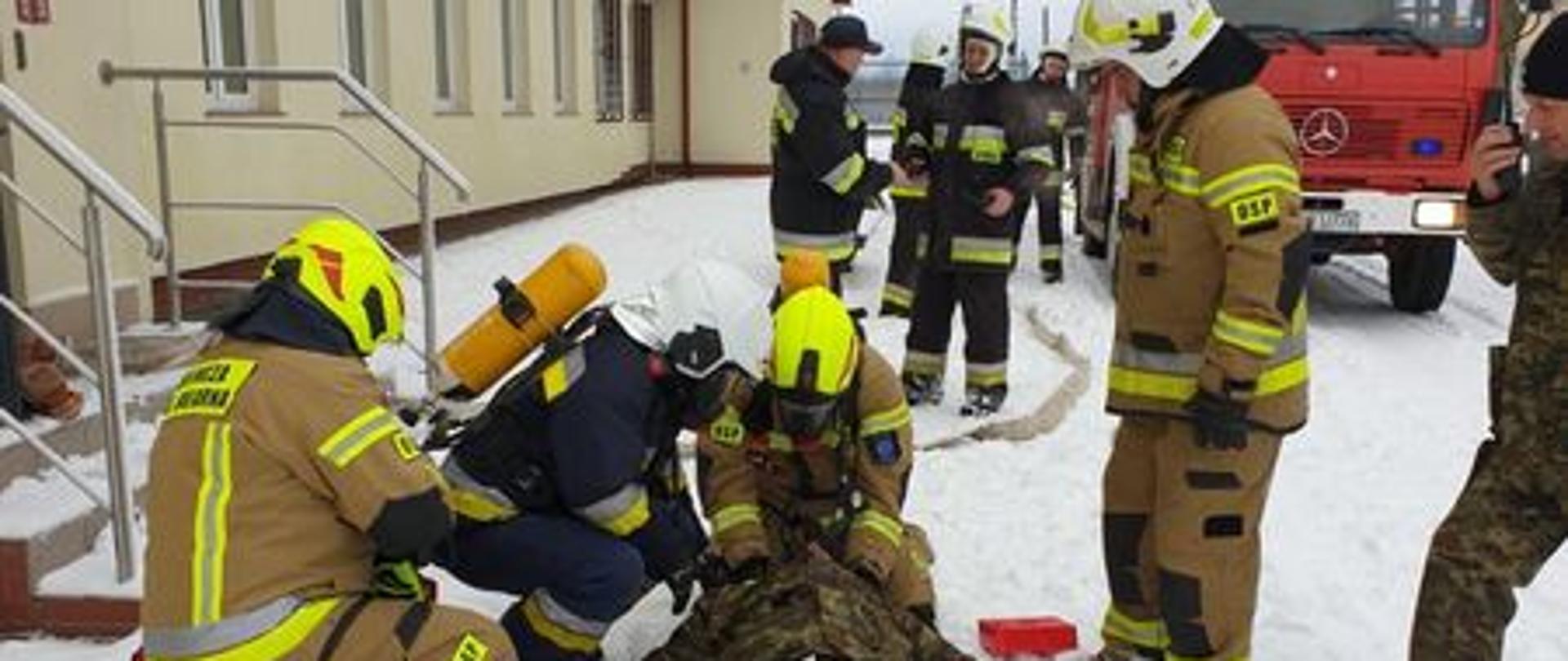 Strażacy i funkcjonariusze w trakcie ćwiczeń przed budynkiem. Jeden leży na śniegu. z prawej strony samochód strażacki
