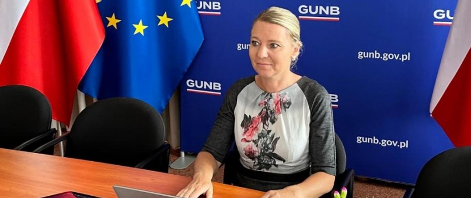 Osoba przy biurku z laptopem, w tle baner GUNB z napisem „gunb.gov.pl”, po bokach flaga Polski i flaga UE.
