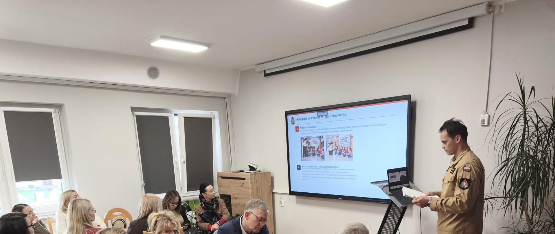 Zdjęcie przedstawia spotkanie w sali konferencyjnej, podczas którego strażak w piaskowym mundurze służbowym stoi przy mównicy i laptopie, omówiąc prezentację wyświetlaną na dużym ekranie ściennym. Slajd nosi tytuł „Kolejność w wyborze miejsc schronienia” i zawiera teksty oraz zdjęcia ilustrujące zasady bezpieczeństwa. Przy długim, drewnianym stole siedzi grupa kilkunastu uczestników spotkania – kobiet i mężczyzn w strojach cywilnych, którzy uważnie słuchają i analizują prezentowane materiały. Po prawej stronie przy ścianie stoi duża roślina doniczkowa, a na podłodze ułożony jest drewniany parkiet.