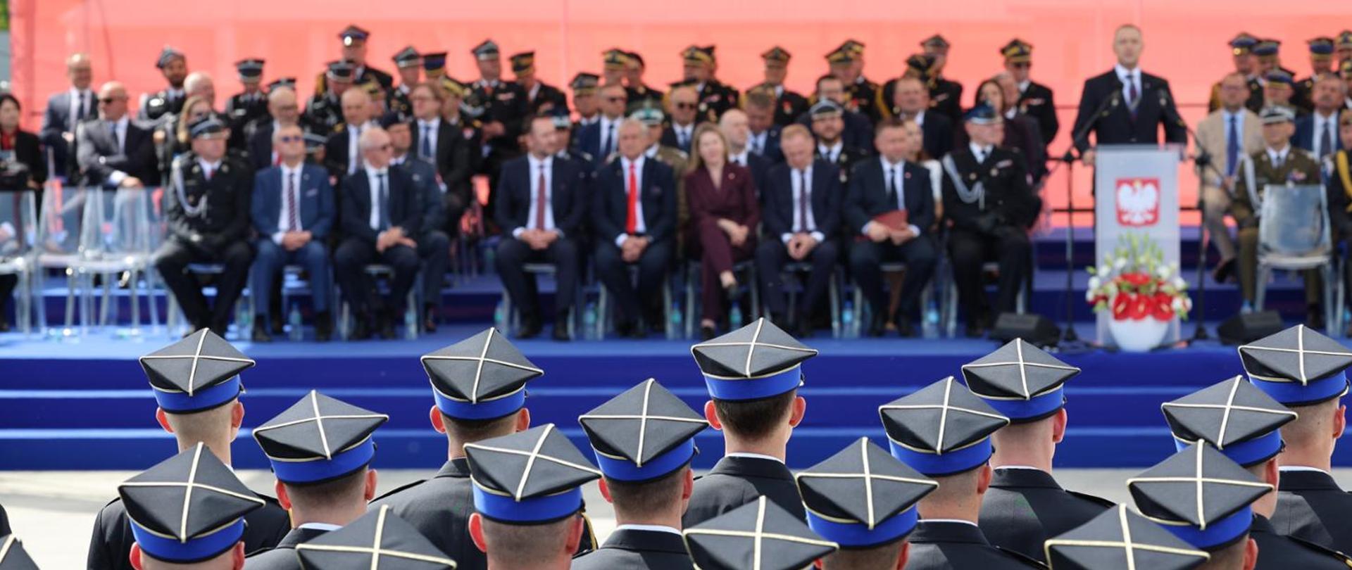 Uroczystość państwowa z udziałem przedstawicieli władz i służb mundurowych. Na pierwszym planie widoczne są tyły głów funkcjonariuszy w charakterystycznych czapkach rogatywkach, skierowanych w stronę trybuny honorowej.
Opis szczegółowy:
Pierwszy plan: Szerokie ujęcie zza pleców grupy funkcjonariuszy (prawdopodobnie Straży Pożarnej lub innej formacji mundurowej). Widoczne są ciemne mundury oraz granatowe czapki rogatywki z niebieskim otokiem i srebrnymi zdobieniami na denku w kształcie krzyża.
Drugi plan: Podwyższona trybuna przykryta niebieską wykładziną. Siedzą na niej zaproszeni goście – mężczyźni w garniturach oraz wysocy rangą oficerowie w galowych mundurach z licznymi odznaczeniami.
Tło: Po prawej stronie zdjęcia znajduje się mównica z godłem Polski (Orłem Białym) na czerwonym tle, przy której stoi przemawiający mężczyzna w ciemnym garniturze. Obok mównicy ustawiony jest kosz z biało-czerwonymi kwiatami. Całość wydarzenia odbywa się na zewnątrz, przy dziennym świetle, na tle czerwonej przesłony w oddali.