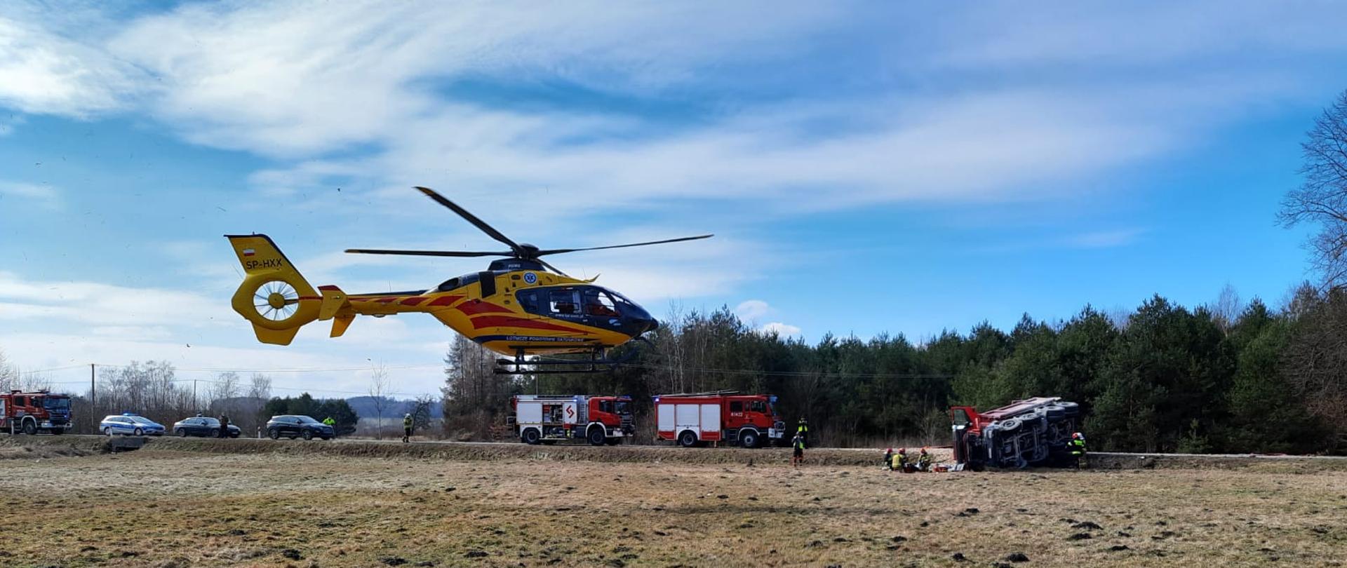 Zdjęcie przedstawia na pierwszym planie po prawej stronie lewy przód pojazdu ratowniczo-gaśniczego GBA 3/16. Z lewej strony widać kabinę przewróconej na bok ciężarówki, z której wyspał się żwir na jezdnię. Na drugim planie, na łące widać śmigłowiec lotniczego pogotowia ratunkowego. Przed kabiną ciężarówki kilku strażaków w ubraniach specjalnych udziela kwalifikowanej pomocy medycznej kierującemu z samochodu ciężarowego. 