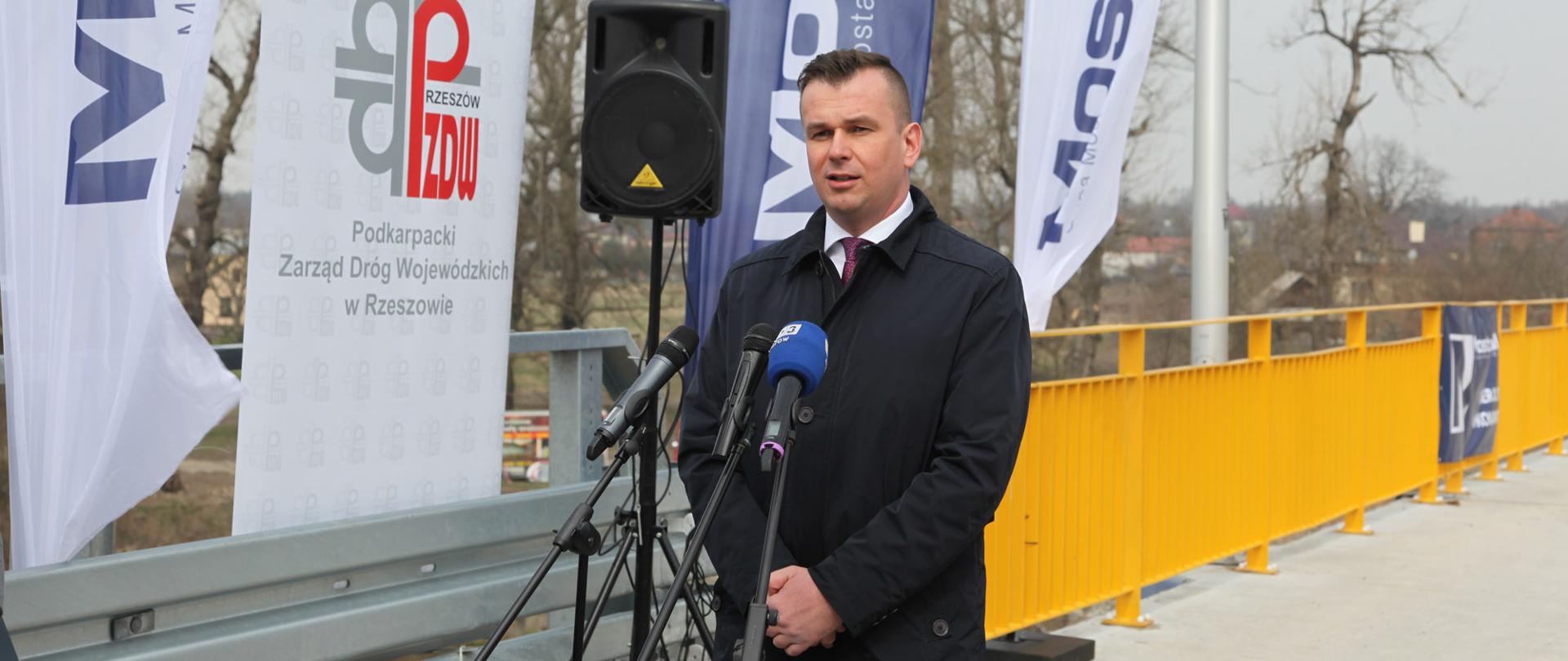 Wiceminister A. Hamryszczak przemawia do mikrofonów na tle mostu.