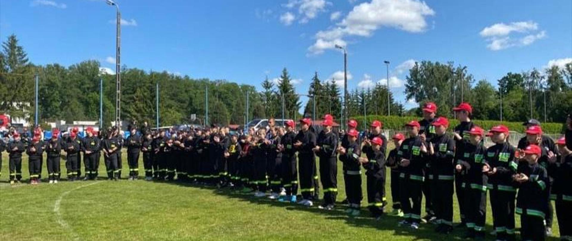 Zbiórka uczestników zawodów młodzieżowych drużyn pożarniczych na boisku miejskiego ośrodka sportu i rekreacji w Rypinie. Zawodnicy ubrani są w ciemne ubrania koszarowe w których występują podczas zawodów. W tle siatka boiska, parking i rosnące w koło drzewa.