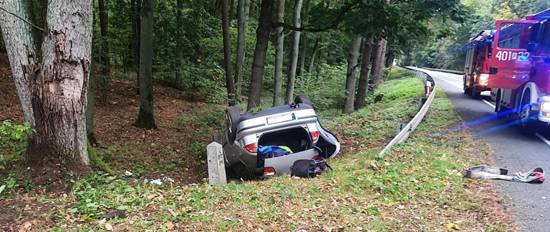 Samochód leży na dachu poza drogą, na której stoją samochody straży pożarnej.