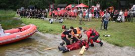 Pokaz z zakresu ratownictwa wodnego wykonany przez ratowników Państwowej Straży Pożarnej oraz Ochotniczej Straży pożarnej w ramach akcji bezpieczne wakacje. Trzech ratowników wydobyło na brzeg jednego ratownika symulującego osobę która się topi. Są zgromadzeni wokół niego nad brzegiem jeziora i przystępują do udzielania mu kwalifikowanej pierwszej pomocy. Za nimi cały pokaz obserwują zgromadzone na plaży dzieci oraz pozostali uczestnicy akcji.