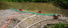 Na zdjęciu widoczny żwirowy brzeg i potok a na nim węże pożarnicze pływające pompy pożarnicze dzięki którym zostaje przepompowywana woda do samochodów pożarniczych.
