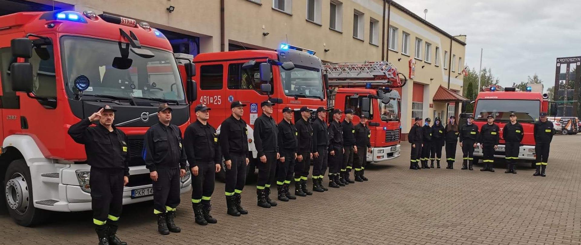 Strażacy na placu przed komendą ustawieni w szyku przed pojazdami pożarniczymi oddają hołd zmarłemu druhowi z jednostki OSP Balice. W pojazdach włączone są sygnały błyskowe. 