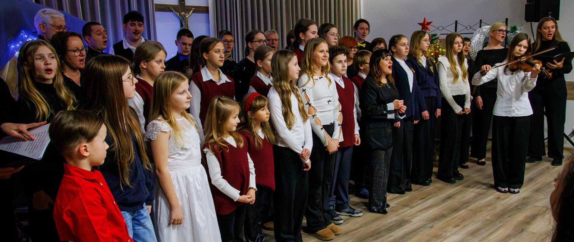 Kilkadziesiąt osób stoi na scenie, osoby śpiewają, jedna z dziewczynek gra na skrzypcach.