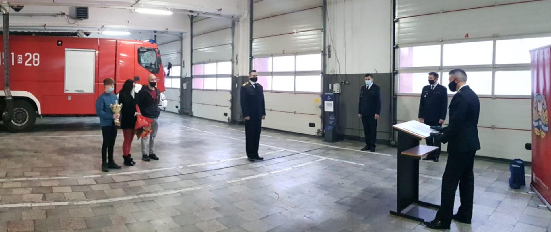 Zdjęcie przedstawia spotkanie na garażu Komendy Miejskiej PSP w Gliwicach. Na zdjęciu znajdują się uratowany chłopiec wraz z rodziną, st. ogn. Składan w mundurze wyjściowym, trzej inni funkcjonariusze w mundurach wyjściowych uczestniczący w spotkaniu, wszyscy uczestnicy maja na twarzach maseczki ochronne, z tyłu w tle stoi czerwony samochód pożarniczy.