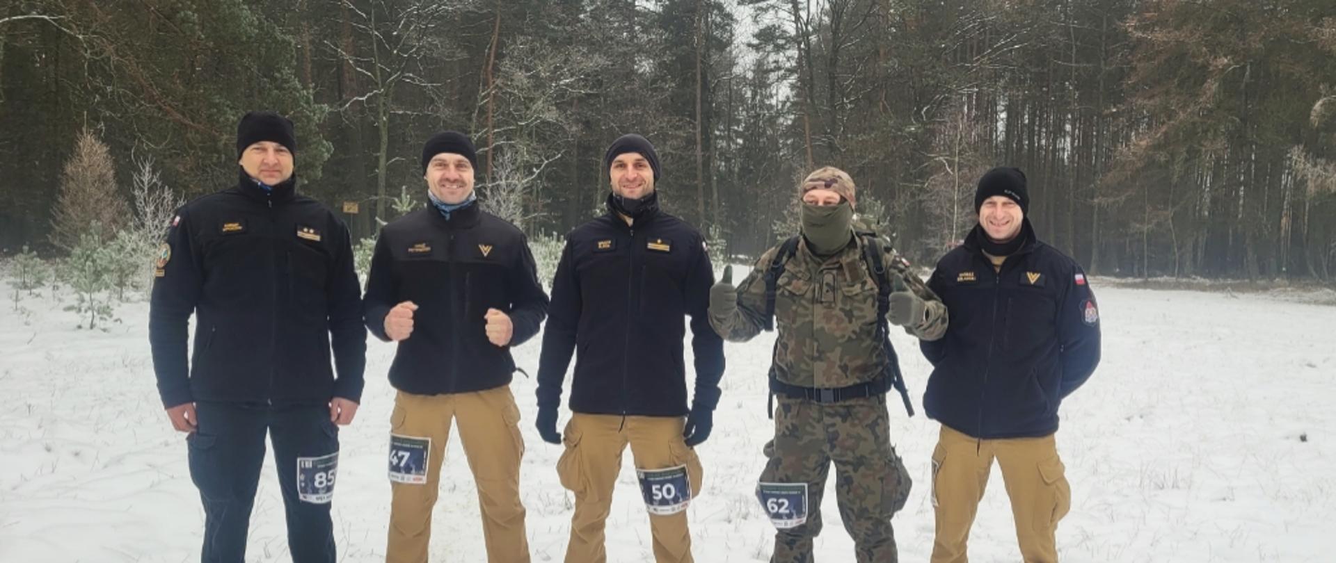 Do zdjęcia pozuje czterech strażaków PSP w czarnych polarach oraz mężczyzna w stroju moro. 