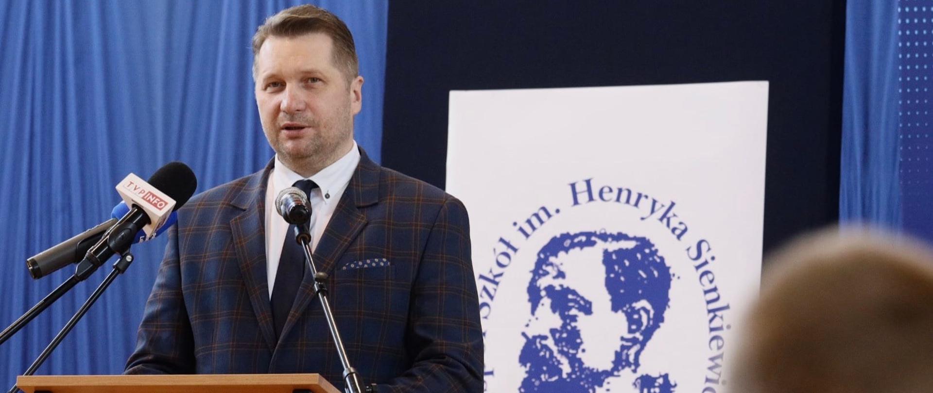Minister Przemysław Czarnek stoi przy mównicy i przemawia, za nim znajduje się tablica z napisem Zespół Szkół im. Henryka Sienkiewicza w Grabowcu.