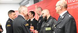 Grupa strażaków w galowych mundurach stoi w szeregu przed czerwonym tłem z logo Państwowej Straży Pożarnej. Jeden z oficerów wręcza odznaczenie w czerwonym etui i uściskiem dłoni gratuluje strażakowi w czarnym ubraniu z naszywką „STRAŻ”. Pozostali strażacy obserwują ceremonię, trzymając w dłoniach medale i odznaczenia.