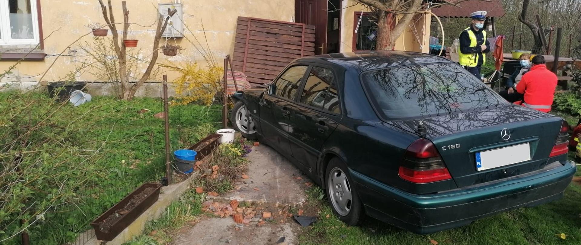 Zdjęcie przedstawia uszkodzony samochód osobowy znajdujący się przed budynkiem mieszkalnym. Po prawej stronie widać funkcjonariusza policji oraz ratownika medycznego, który udziela pomocy starszej kobiecie.