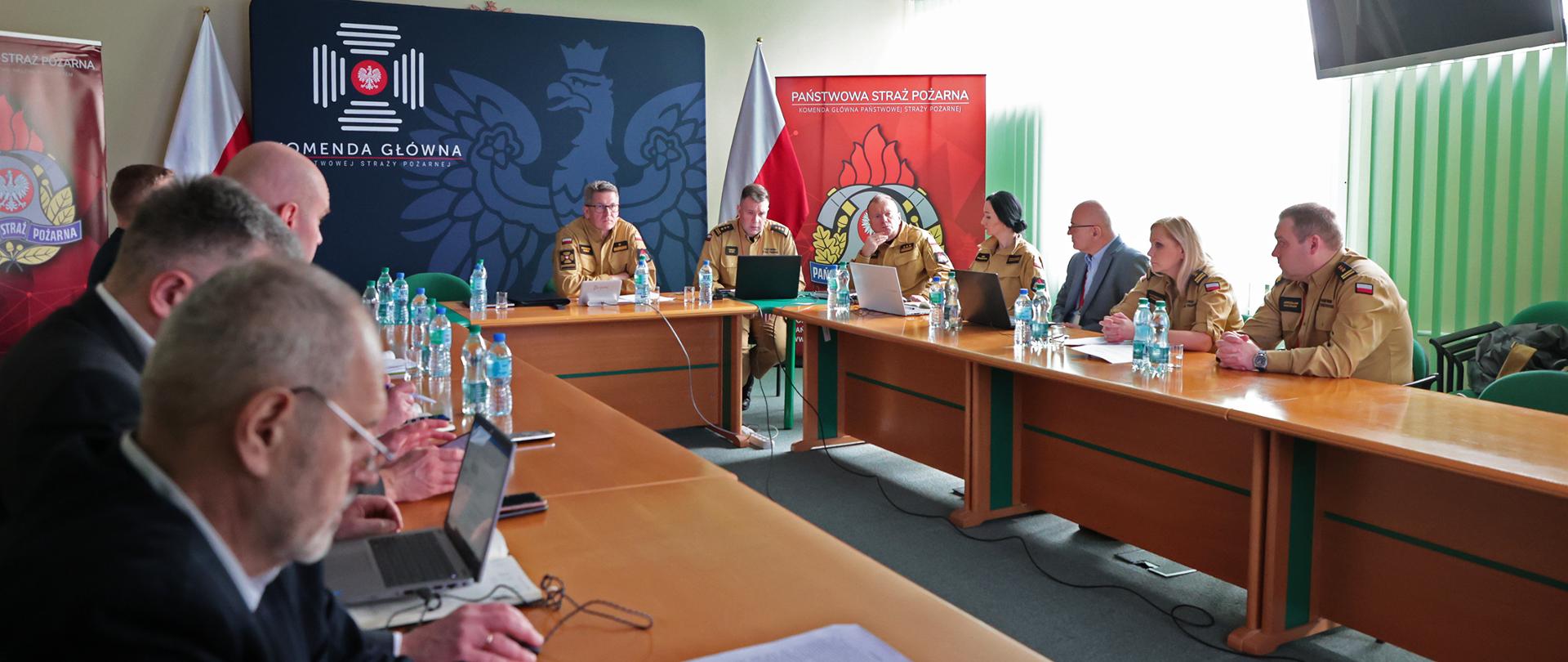 Widok ogólny sali na której przy stołach siedzi kilku mężczyzn oraz dwie kobiety w piaskowych mundurach strażackich. Na stole ustawionym w podkowę stoją butelki z wodą, laptopy oraz dokumenty 