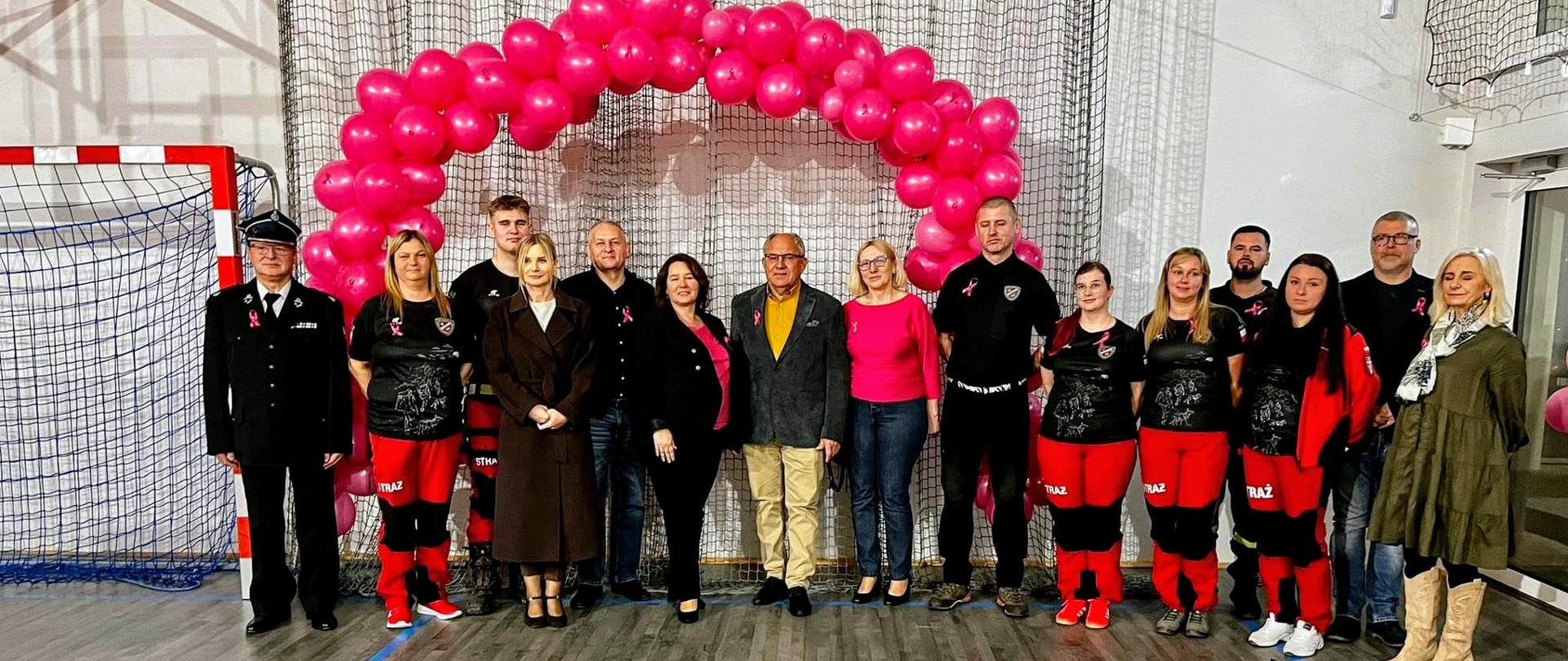 Na zdjęciu grupa kilkunastu osób stoi w rzędzie w sali gimnastycznej. Za nimi widać duży łuk z różowych balonów oraz siatkę ochronną na ścianie. Część osób ubrana jest w czarne i czerwone stroje strażackie z napisem „STRAŻ”, inni w eleganckie ubrania cywilne. Na podłodze przed grupą leżą różne torby i sprzęt medyczny, w tym butla z tlenem, nosze, apteczki i zestawy opatrunkowe.