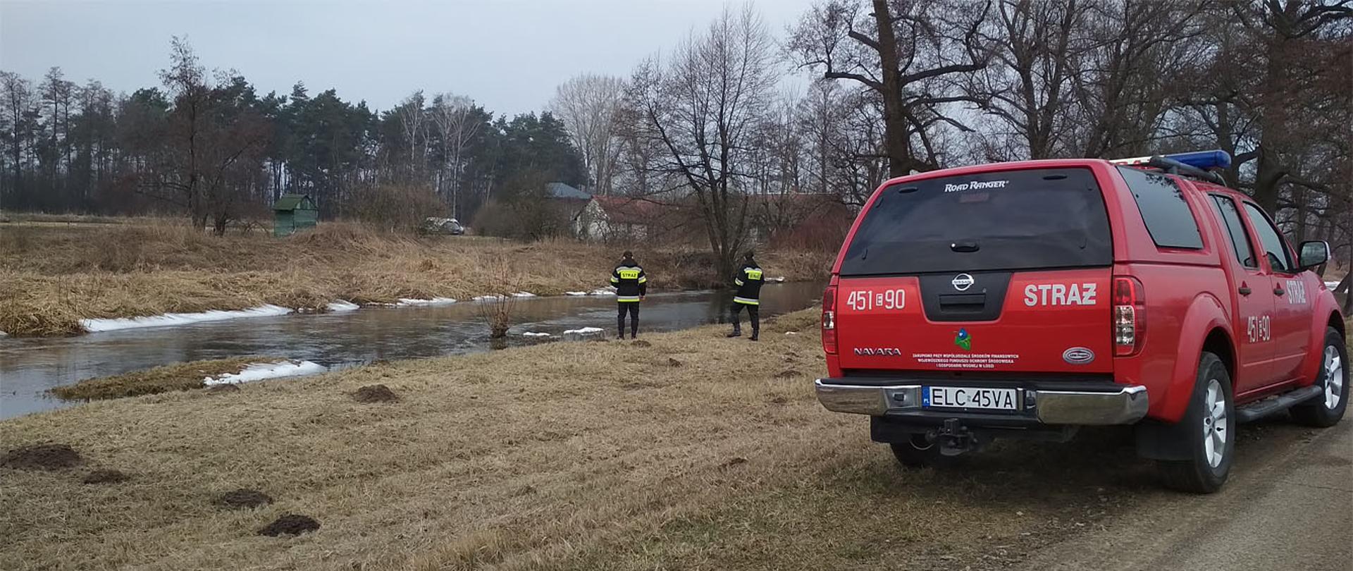 Zdjęcie przedstawia dwóch strażaków ubranych w ubranie sztabowe barwy czarnej z odblaskami koloru szarego i jasnożółtego z nakryciem głowy w postaci czapki z daszkiem oglądających poziom wody w rzece Mroga w miejscowości Bielawy. W tle zabudowa mieszkalna w otoczeniu drzew. Na pierwszy planie widać samochód strażacki typu terenowego barwy czerwonej z numerem operacyjnym 451E90 oraz napisem STRAŻ. Fotografia przedstawia resztki topniejącego śniegu, widoczna jest mocno pożółkła krótka trawa.