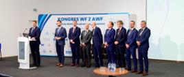 Na sali pod ścianą z napisem Kongres WF z AWF stoi grupa mężczyzn w garniturach, przed nimi minister Czarnek stoi za mównicą i mówi do mikrofonu.