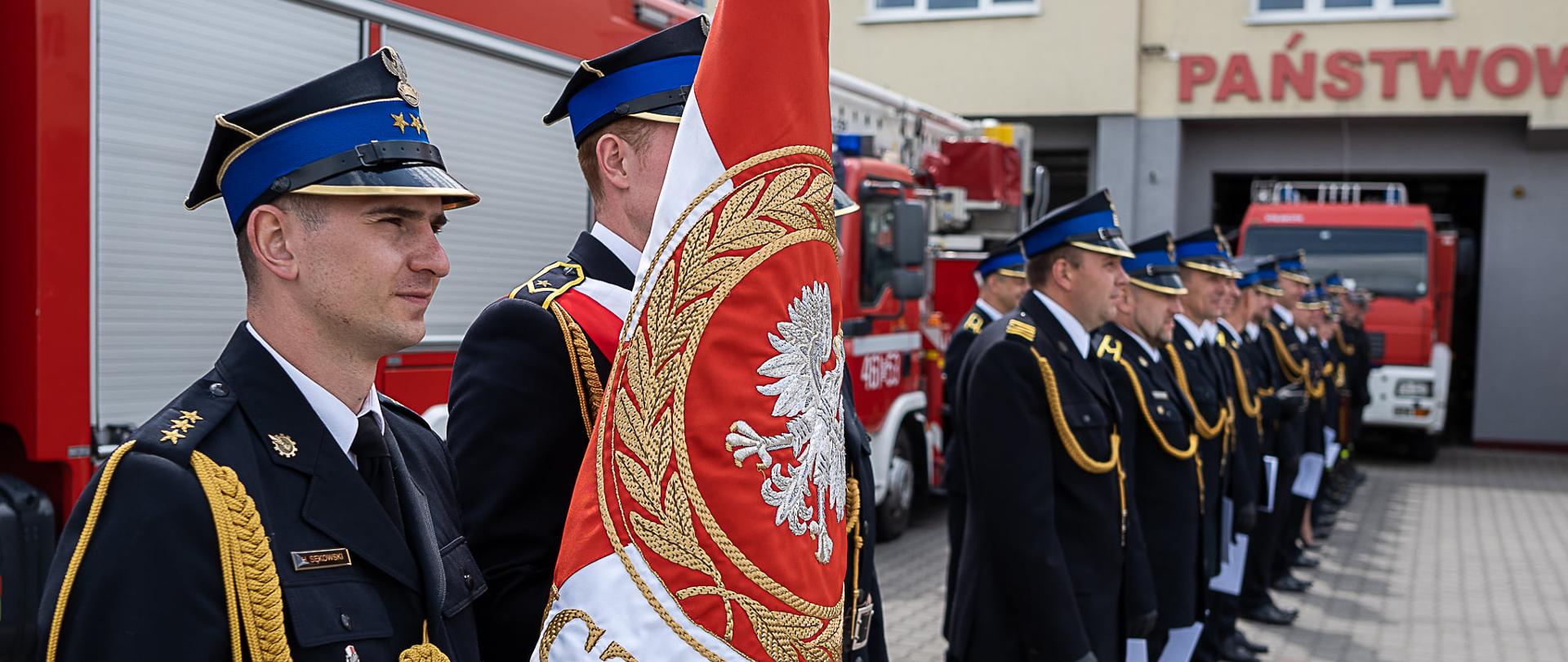 Zdjęcie przestawia poczet sztandarowy Komendy Powiatowej PSP powiatu łódzkiego wschodniego, na dalszym planie widać pododdziały podczas uroczystego apelu. 