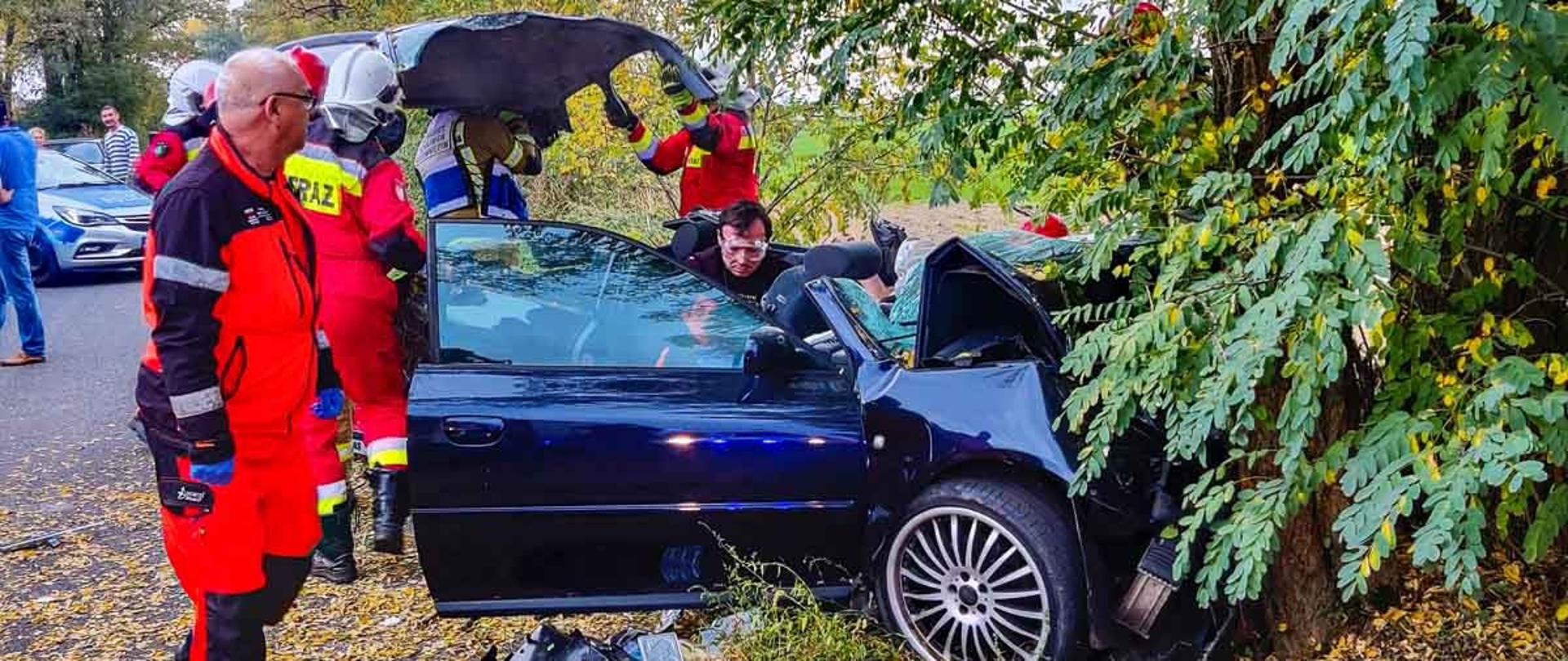 Na zdjęciu granatowy samochód osobowy który uderzył w drzewo rozbity, obok strażacy zdejmujący dach samochodu który został rozcięty przy aucie i w pojeździe ratownicy medyczni