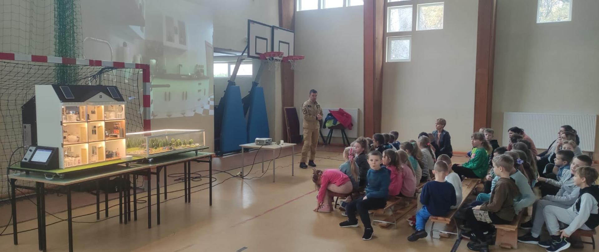 Strażak w mundurze koloru musztardowego trzyma mikrofon opowiadając dzieciom o zagrożeniach mogących wystąpić w domu na przykładzie symulatora zagrożeń przeciwpożarowych obok siedzi pani nauczyciel. 