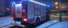 Panuje zmrok, pada intensywnie śnieg, na drodze przed domem stoi oświetlony samochód ratowniczo - gaśniczy obok niego strażak w umundurowaniu bojowym