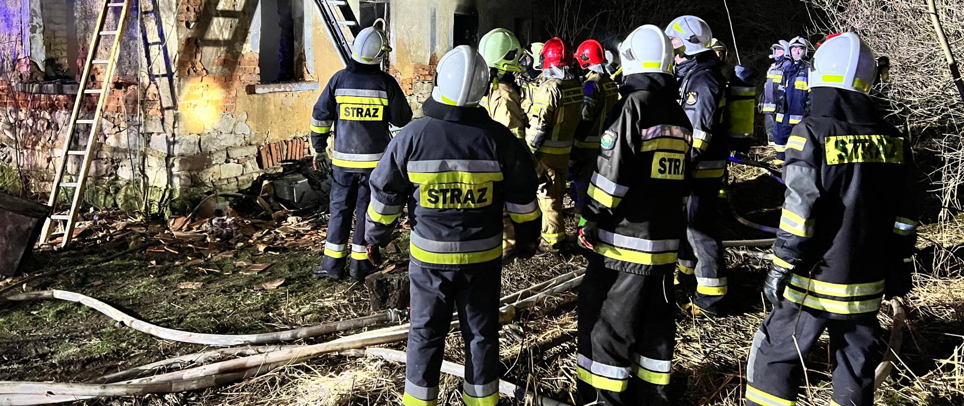 dom jednorodzinny z którego wydobywa się dym, wokół strażacy oraz dwie drabiny przystawione do budynku
