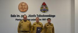 Trzech strażaków Państwowej Straży Pożarnej stoi obok siebie funkcjonariusz stojący w środku trzyma czarną teczkę z orzełkiem na ścianie wisi logo Państwowej Straży Pożarnej obraz patrona sali oraz napis sala imieniem podpułkownika Józefa Tuliszkowskiego pioniera polskiego pożarnictwa tysiąć osiemset sześćdziesiąt siedem myślnik.