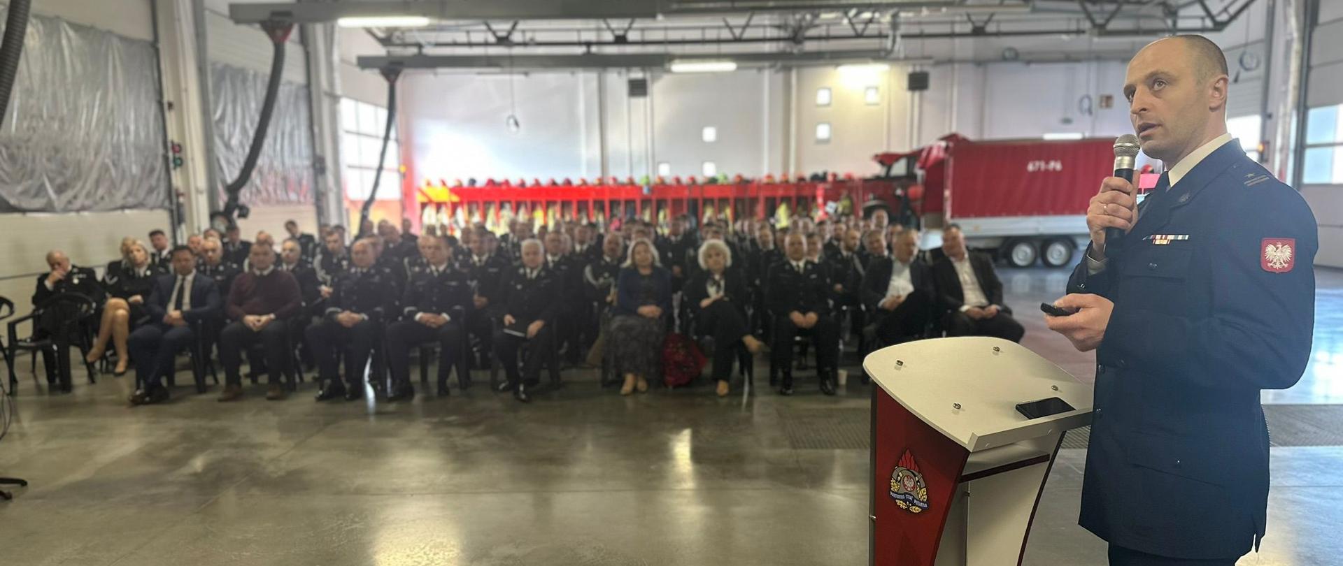 Na zdjęciu widać strażaka w mundurze wyjściowym (Komendant KP PSP Węgrów), który przemawia, przed zgromadzonymi gośćmi. Odprawa odbywa się w garażu siedziby KP PSP Węgrów.