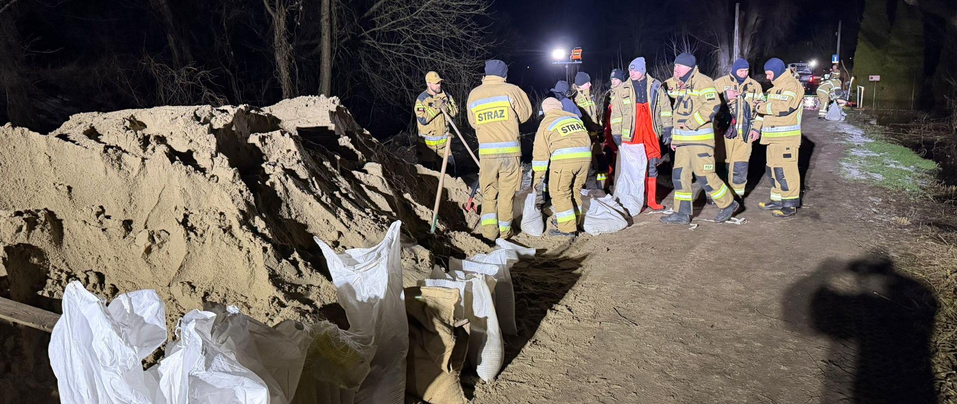 Strażacy ochotnicy i funkcjonariusze służb ratowniczych w nocy napełniają i układają worki z piaskiem na drodze w pobliżu rzeki. Obok znajduje się duża hałda piasku oraz rząd przygotowanych worków. Działania prowadzone są przy sztucznym oświetleniu i mają na celu zabezpieczenie terenu przed skutkami podwyższonego poziomu wody