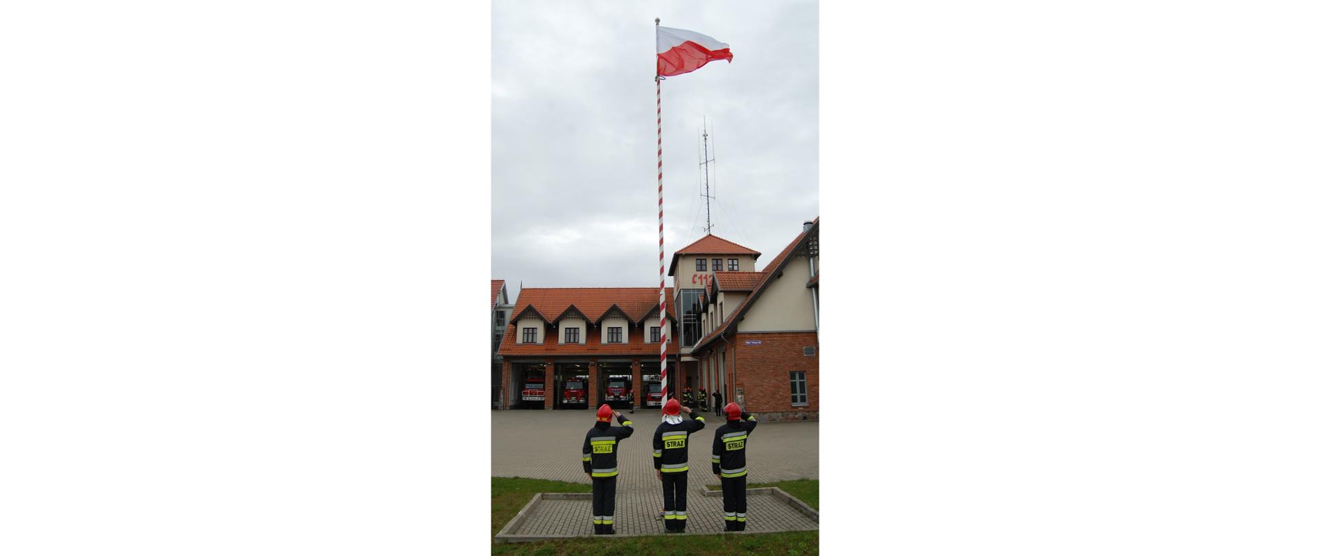 Zdjęcie przedstawia funkcjonariuszy KP PSP w Gołdapi uczestniczących podczas uroczystej zmiany służby z okazji Dnia Flagi Rzeczypospolitej Polskiej, oddających honor w momencie podnoszenia flagi RP na maszt i akompaniamencie hymnu państwowego