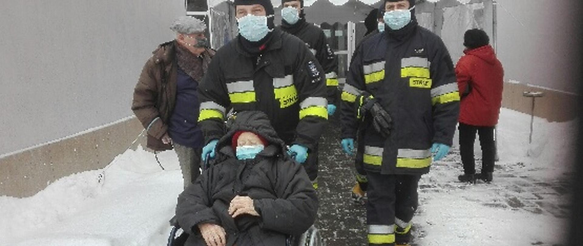 Obraz przedstawia strażaków w umundurowaniu specjalnym z założonymi maseczkami ochronnymi na twarzy. Strażacy prowadzą wózek inwalidzki ze starszą osobą. W tle budynek przychodni i zaśnieżony chodnik.