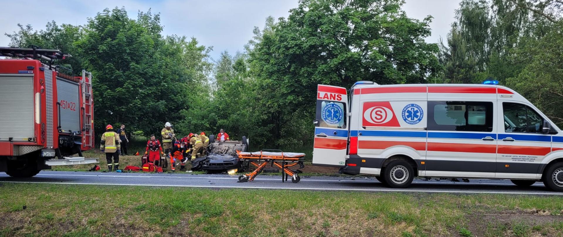 Zdjęcie wykonane z oddali. Widać strażaków i ratowników pogotowia ratunkowego udzielający pomoc jednemu z kierowcy samochodu audi, który wyniku zderzenia czołowego z drugim samochodem dachował.