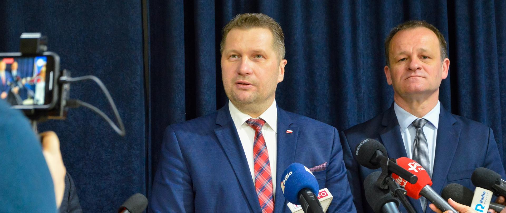 Na tle przysłoniętej granatowym materiałem ściany stoi minister Czarnek który mówi do kilku mikrofonów na stojakach, przed nim dziennikach trzymający mikrofon, za ministrem stoi mężczyzna w czarnym garniturze,