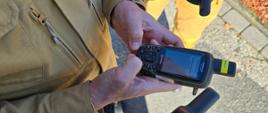 3. Na zdjęciu jest zbliżenie na trzy dłonie trzymające trzy różne ręczne odbiorniki GPS marki Garmin, sugerując, że osoby konfigurują urządzenia. W centrum uwagi znajdują się dłonie (jedna w rękawie jasnego, beżowego munduru – prawdopodobnie strażaka z PSP) trzymające dwa urządzenia. W tle widać fragmenty beżowych spodni. Zdjęcie dokumentuje szkolenie z wykorzystaniem sprzętu nawigacyjnego.