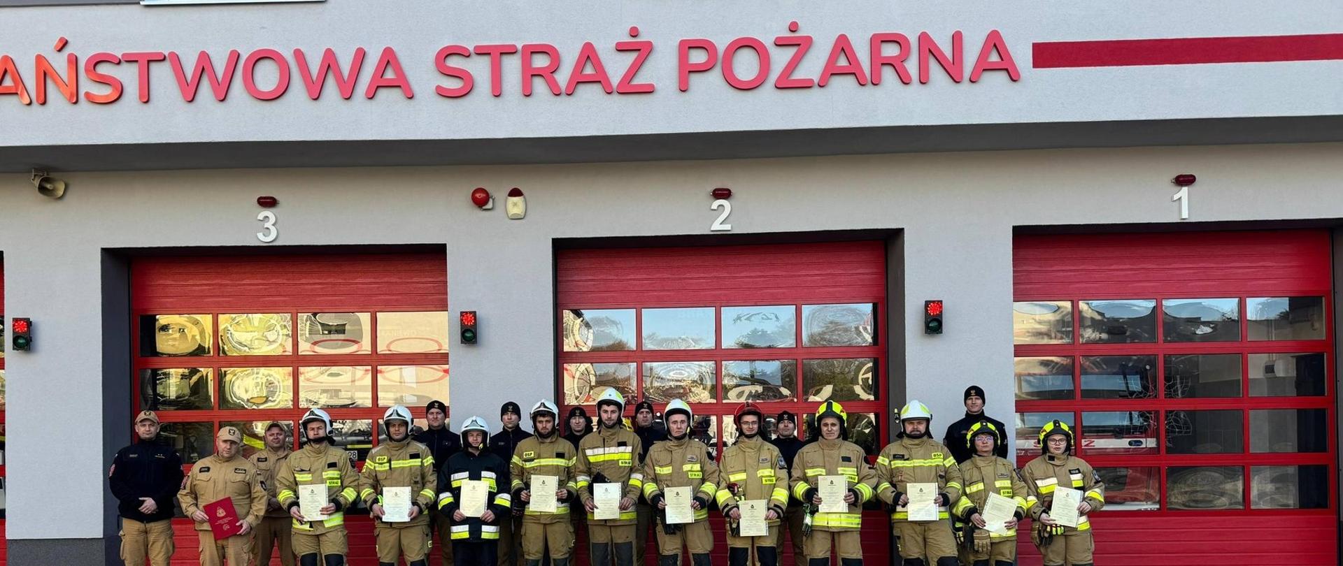 Na tle czerwonych bram garażu bojowego państwowej straży pożarnej stoją uczestnicy szkolenie, strażacy OSP w mundurach bojowych i hełmach.