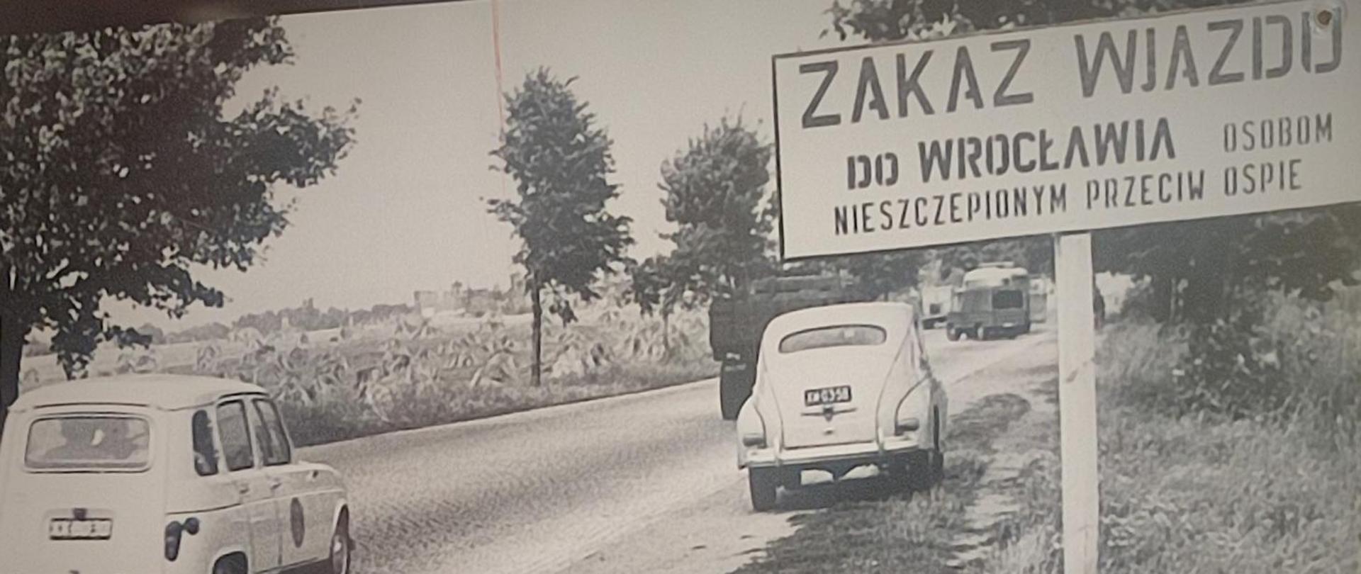 Czarno-białe zdjęcie historyczne przedstawiające najprawdopodobniej drogę wjazdową do Wrocławia. Po prawej stronie drogi widać znak z napisem: "ZAKAZ WJAZDU DO WROCŁAWIA OSOBOM NIESZCEPIONYM PRZECIW OSPIE".