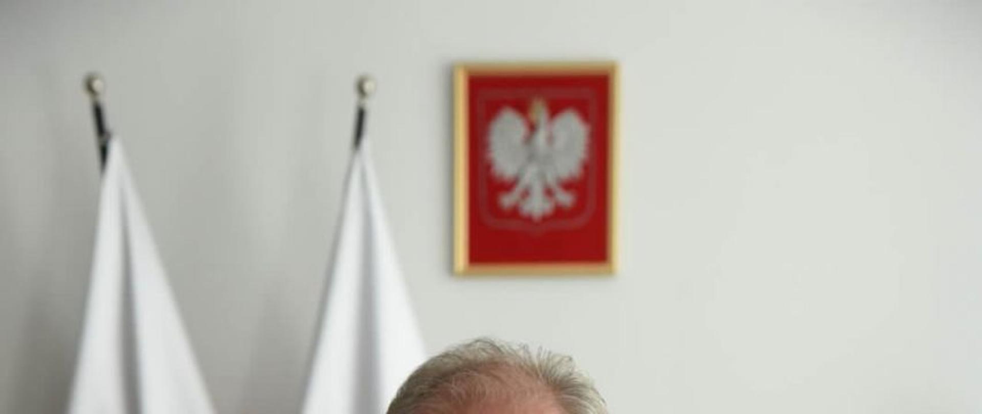 na zdjęciu przy biurku siedzi wojewoda pomorski podpisujący dokumenty, w tle stoją dwie flagi państwowe, na ścianie wisi godło państwowe.