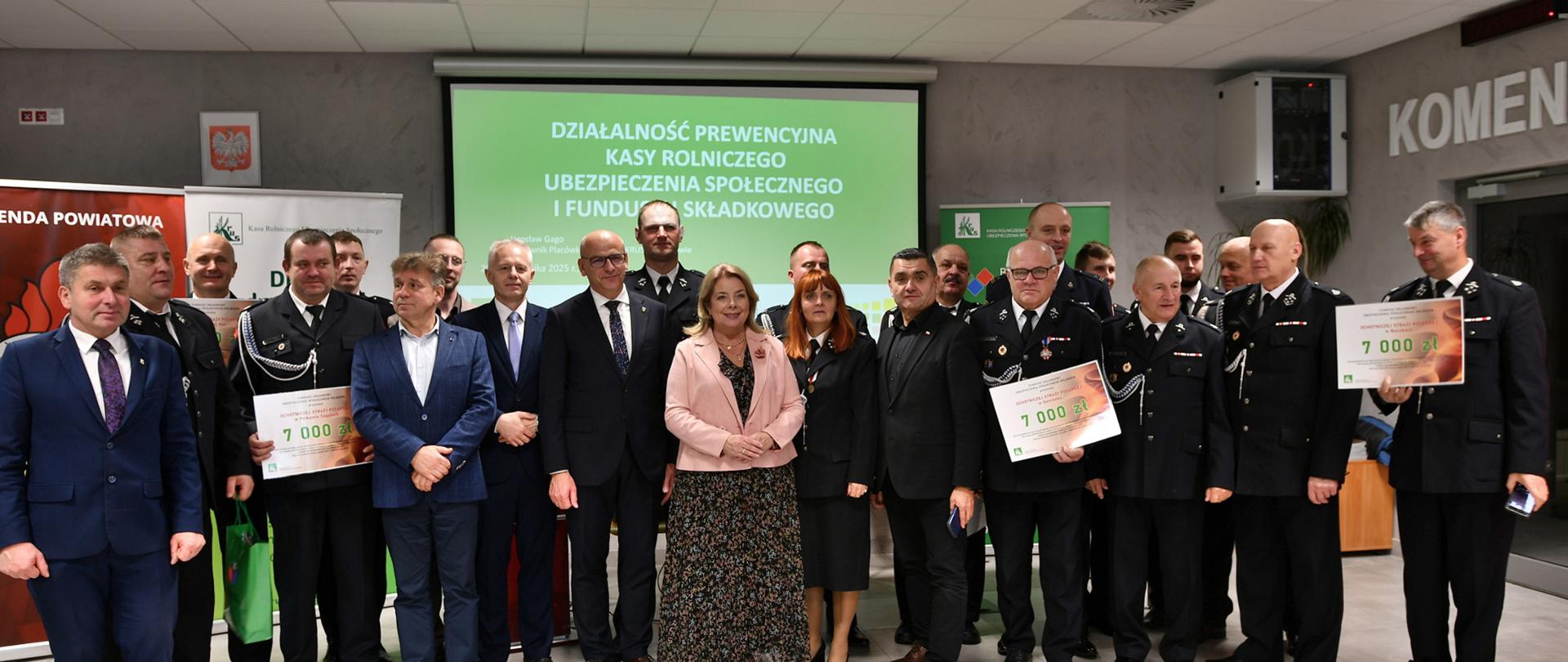 Na tle w slajdu prezentacji i banerów stoi duża grupa mężczyzn, na środku dwie kobiety, kilku mężczyzn trzyma w rękach duże vouchery. 