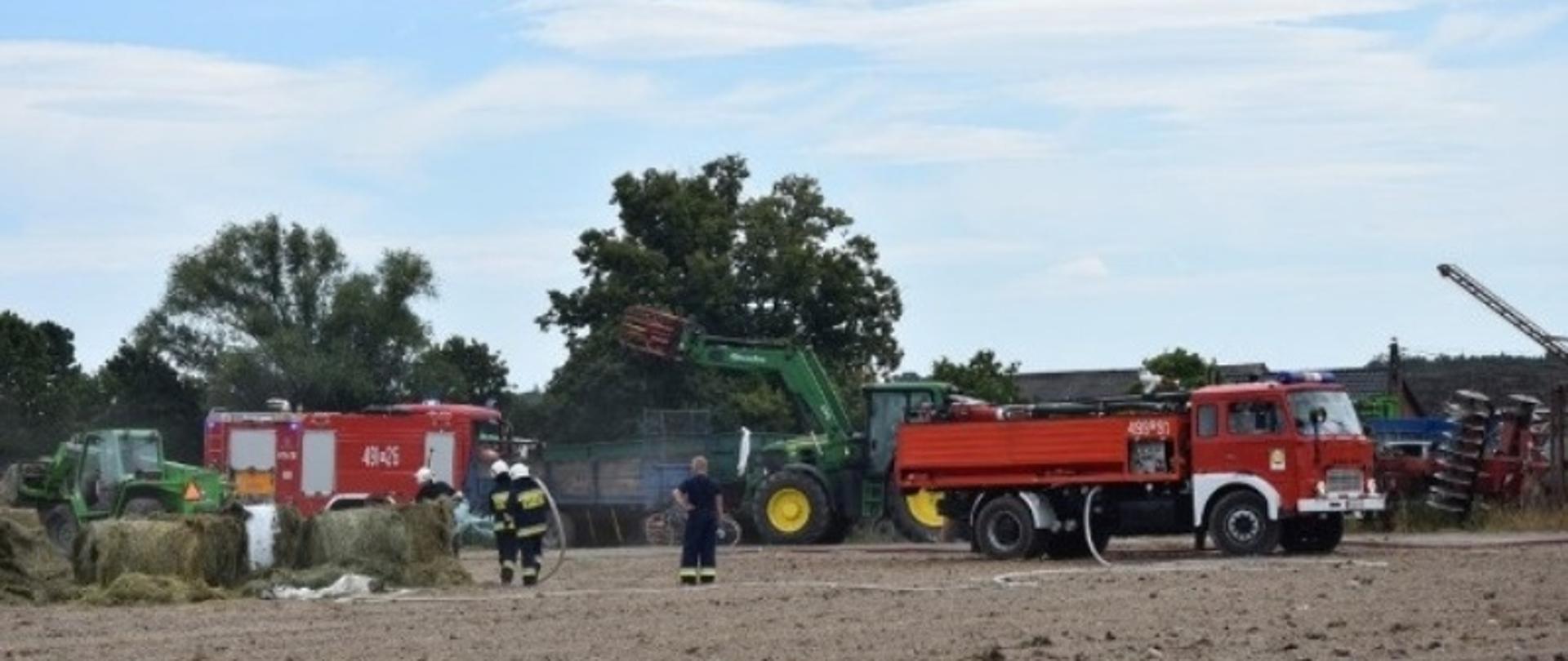 Dogaszanie tlących się balotów na pobliskim polu. Dwóch strażaków w ubraniach bojowych trzyma odcinek węża i polewa wodą tlące się baloty, które wywozi sprzęt rolniczy. Dwóch kolejnych strażaków za pomocą wideł rozgarnia baloty celem dogaszenia zarzewi ognia. Zasilanie linii gaśniczej realizowane jest z samochodu ciężkiego typu jelcz. W oddali widać maszyny rolnicze na placu gospodarstwa. 