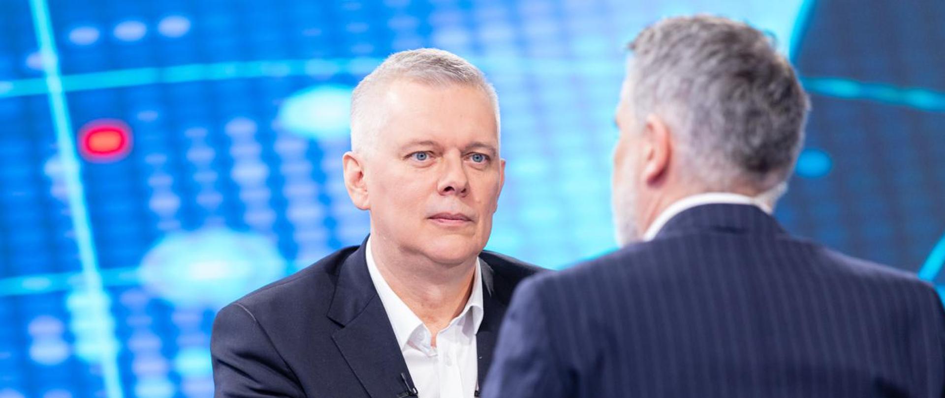 Na zdjęciu dwie siedzące przy stole osoby - po lewej minister Tomasz Siemoniak, a po prawej (siedzący tyłem) redaktor Mariusz Piekarski. Na stole leżą materiały. W tle widać niebiesko-seledynowy ekran. 